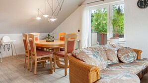 A dining area with a wooden table and chairs, a wicker sofa, and a large window with a view of greenery. - Apartment in Bad Wunnenberg Near Ski Willingen (Bad Wuennenberg)
