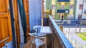 Un balcon avec une table et deux chaises, une porte en bois et une vue sur un immeuble verdoyant de l’autre côté de la rue.