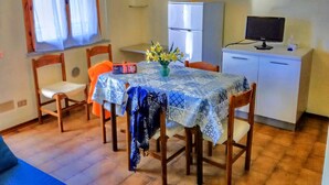 Un petit coin repas avec une table recouverte d’une nappe bleue à motifs, entourée de chaises en bois. Un réfrigérateur et un téléviseur sont posés sur un meuble, à droite.