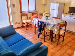 A dining area with a table covered by a patterned tablecloth, surrounded by wooden chairs. A blue sofa is in the foreground, and a refrigerator is visible in the background. - Apartment 200m From the sea (Ravenna)