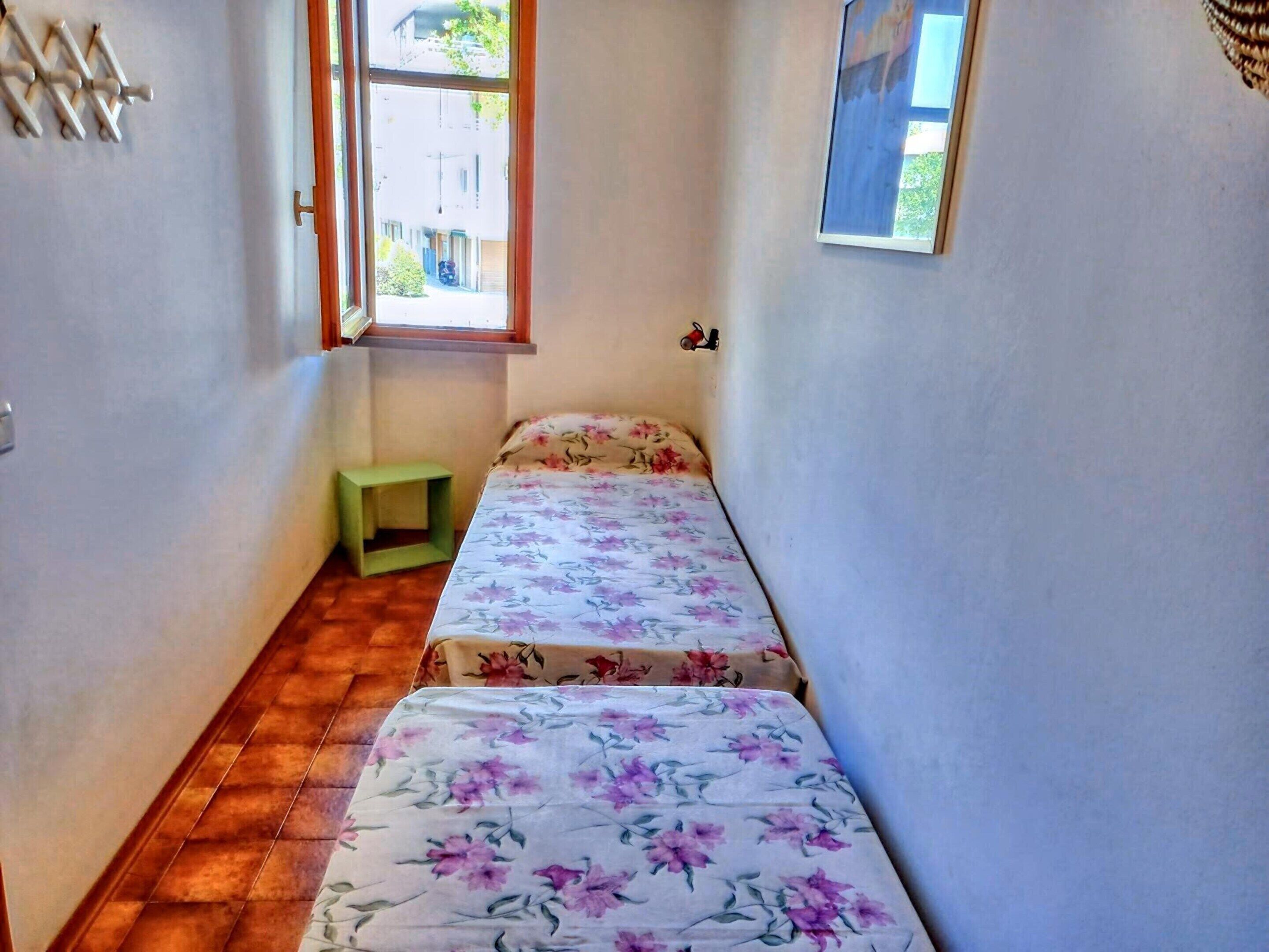 A narrow room with two single beds, a small green nightstand, a window with a view of buildings, and a wall-mounted light fixture.