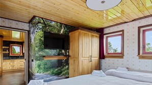 Quarto com cama, guarda-roupa de madeira, televisão na parede e janela com vista para árvores.