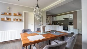 Un comedor moderno con una mesa de madera, sillas tapizadas en gris, una araña y una cocina al fondo.