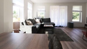 A modern living room with a sectional sofa, a coffee table, and a dining table. There are large windows with sheer curtains, a lamp, and a TV stand. - Holiday Home in South Holland Near Rotterdam (Simonshaven)