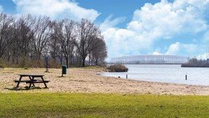 A park with a picnic table and a bench, a sandy area near a body of water, and leafless trees in the background. - Holiday Home in South Holland Near Rotterdam (Simonshaven)