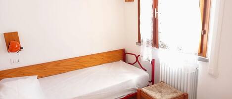 Una cama individual con cabecera de madera, un taburete pequeño de madera, una lámpara fijada a la pared y una ventana con cortinas traslúcidas.