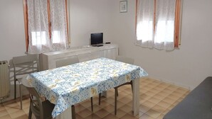 A small dining area with a table covered in a patterned tablecloth, two chairs, a television on a white cabinet, and two windows with curtains. - Cosy Apartment With Balcony (Ravenna)