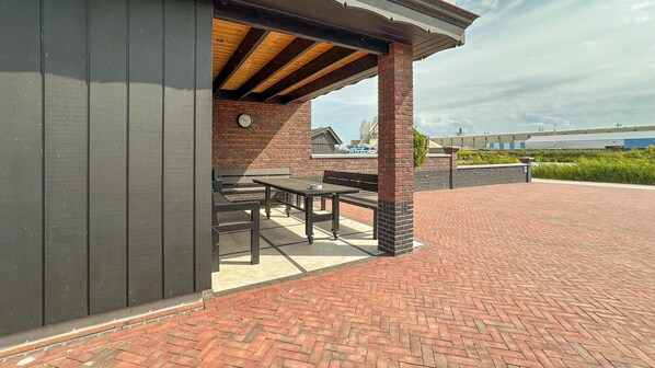 Une terrasse extérieure avec un sol en briques et un espace salon couvert, équipé de tables et de bancs.