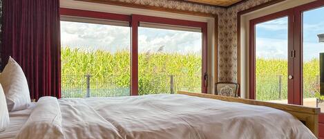 Une chambre avec un grand lit, une vue sur un champ de maïs par une porte coulissante et un balcon avec une chaise.