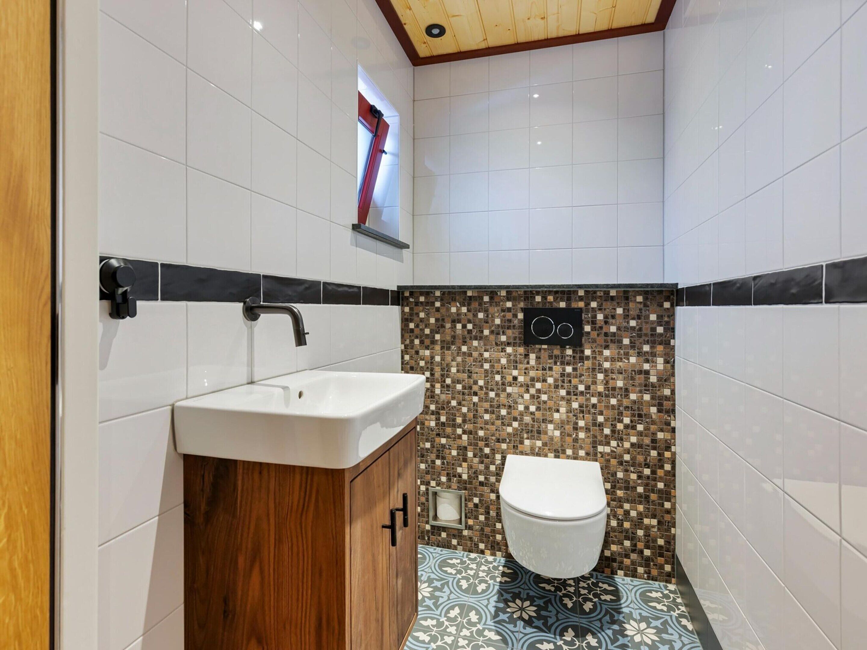 A modern bathroom with white tiles, a wooden vanity, a toilet, and a mosaic-patterned wall.