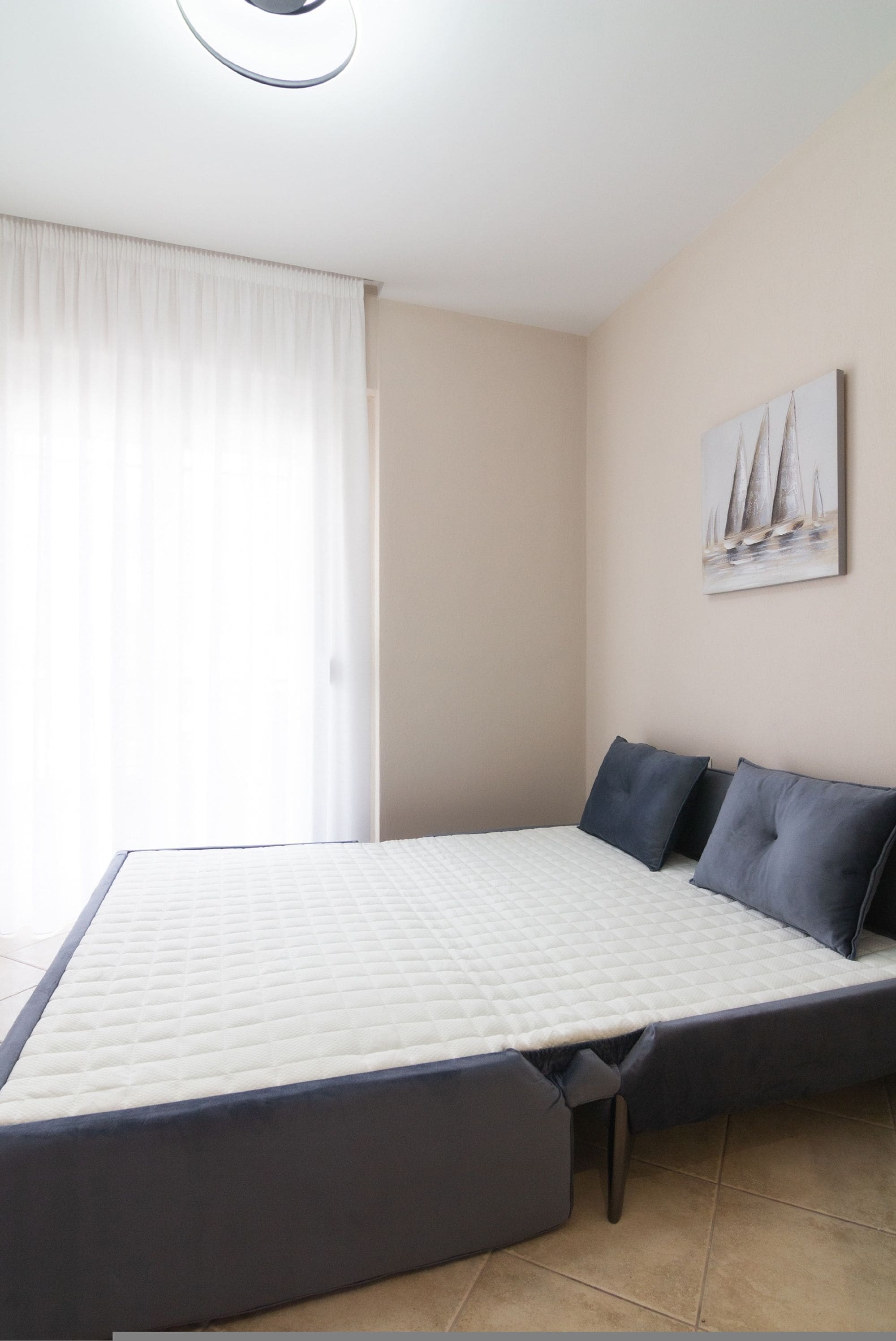 Un dormitorio con una cama, una ventana con cortinas, un cuadro colgado en la pared y suelo de baldosas.