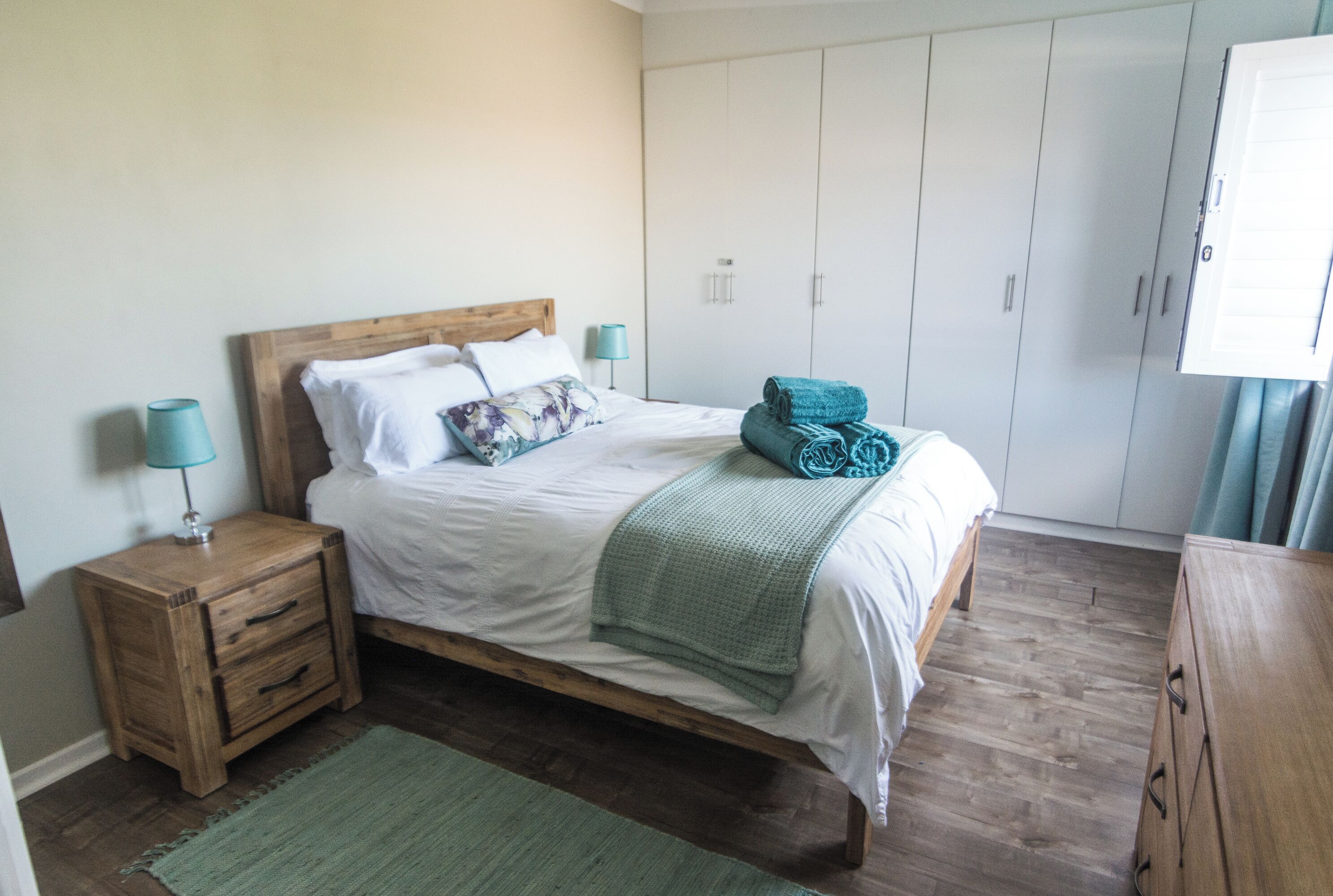 Un lit bien fait, avec une tête de lit en bois, une couverture verte et un coussin assorti. Une table de chevet en bois avec une lampe se trouve à côté du lit. Un tapis vert est posé sur le sol, et une armoire blanche se trouve à l’arrière-plan.