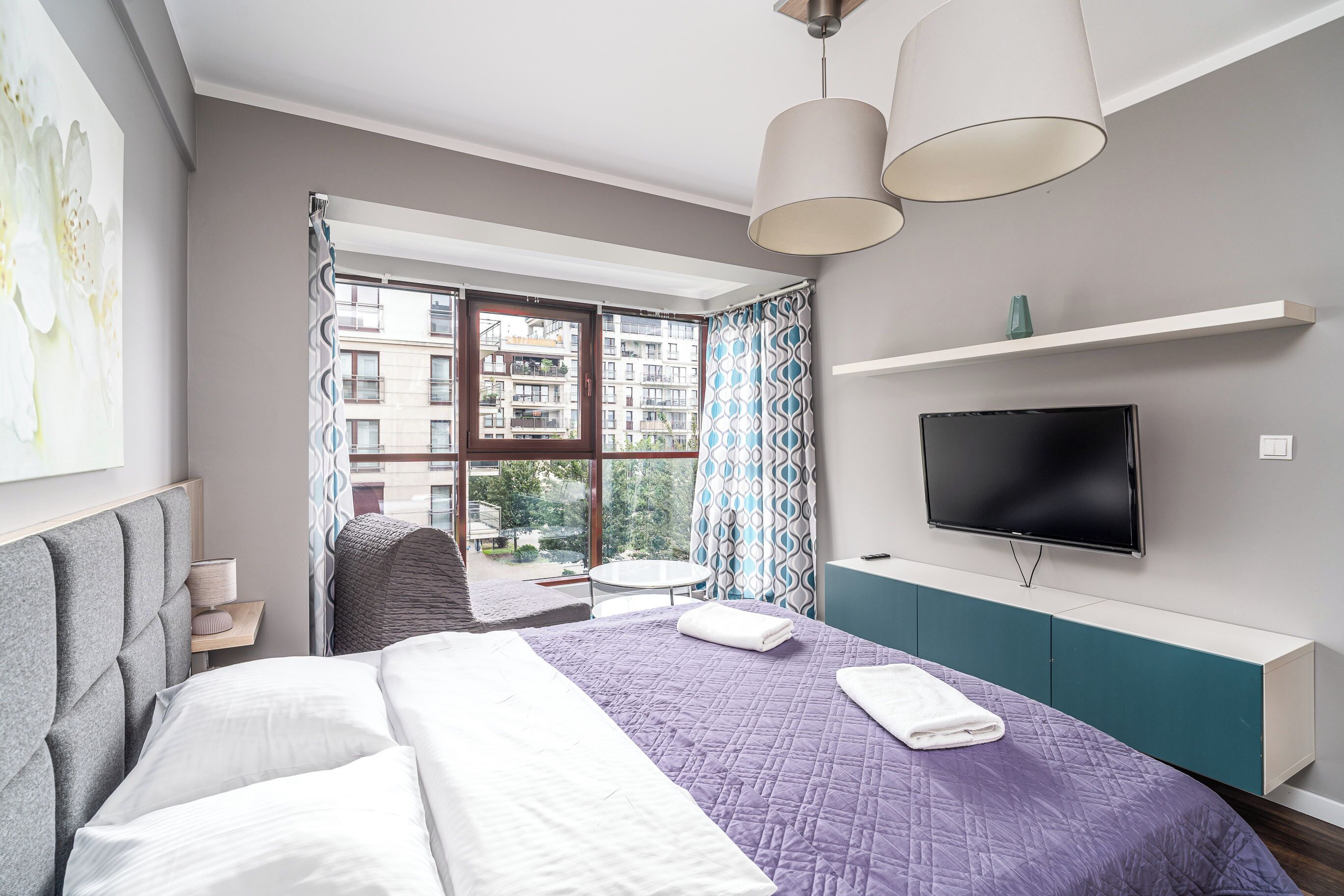 A modern hotel room with a large bed, a TV, a window with a view of buildings, and a balcony.