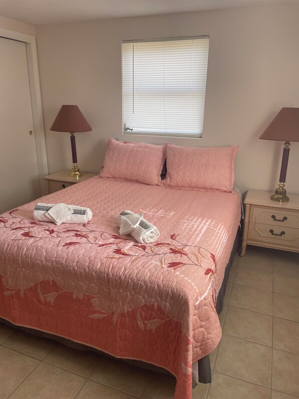 Une chambre à coucher avec une couvre-lit rose matelassé, deux lampes de chevet, une fenêtre avec des stores et une porte.