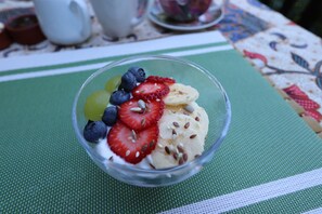 En genomskinlig glasskål med skivade jordgubbar, bananer och blåbär, placerad på en grön bordsduk.