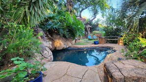 A small, naturalistic pool surrounded by rocks and lush vegetation, with a wooden deck and a stone pathway leading to a gazebo.
