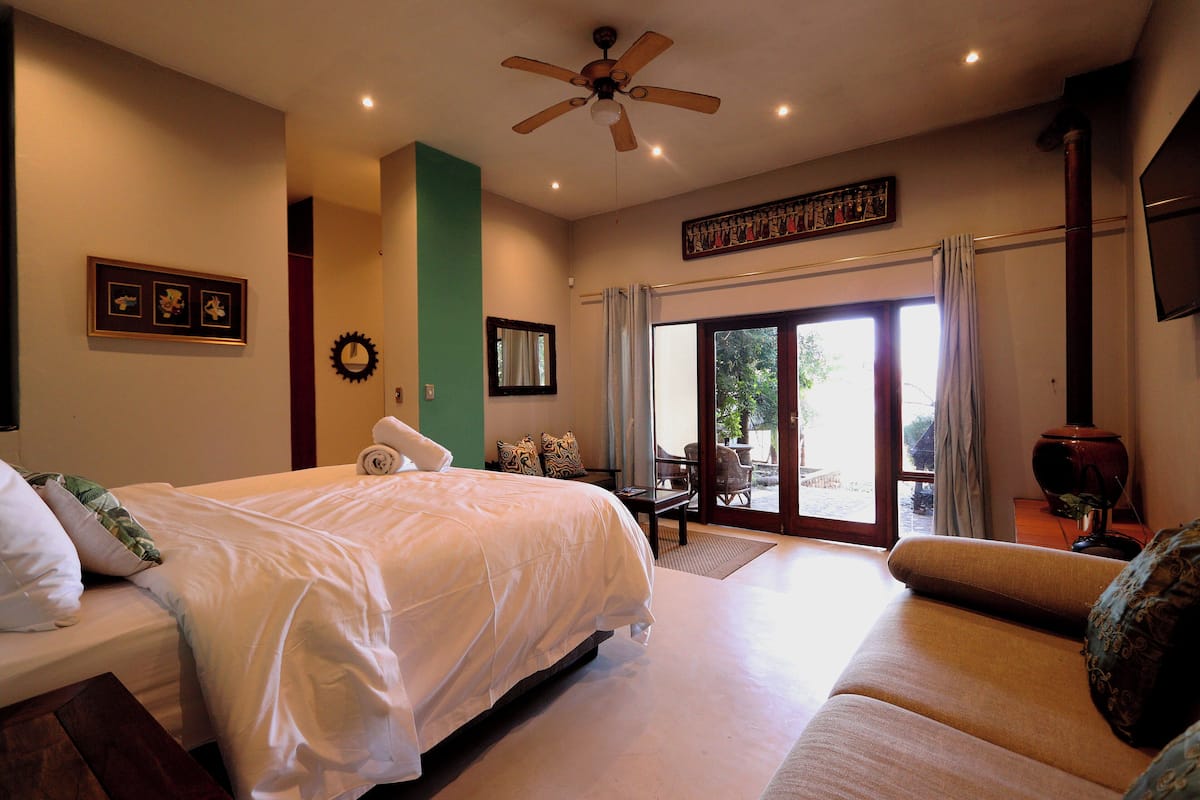 Habitación de hotel con una cama grande, un sofá, un ventilador de techo, una mesa de comedor y vistas al exterior a través de la puerta de cristal.