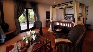 A bedroom with a four-poster bed, a sitting area with wicker chairs, a glass coffee table with a vase, and a view of the outdoors through a glass door.
