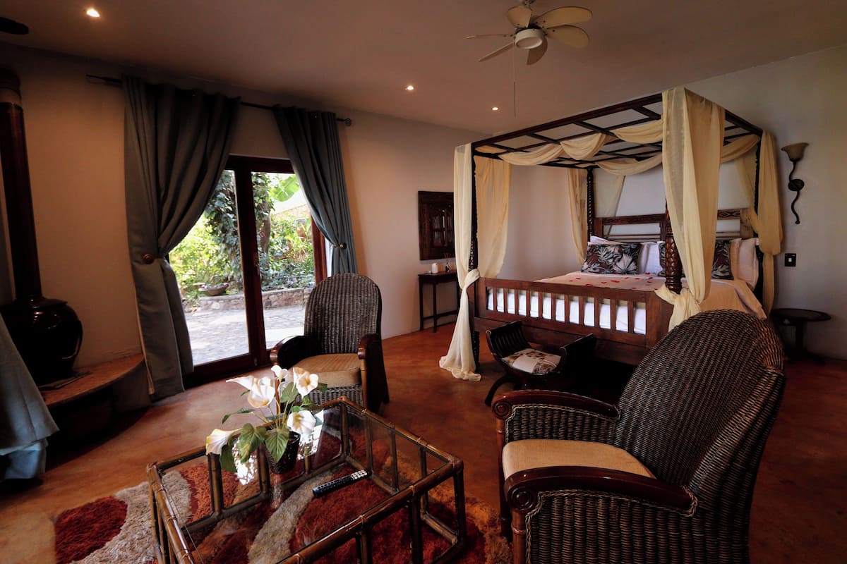 Un dormitorio con cama con dosel, una zona de estar con sillones de mimbre, una mesa de centro de vidrio con un florero, y vista al exterior a través de una puerta de cristal.