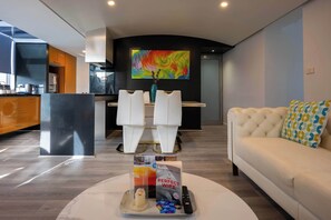 Un soggiorno moderno con un divano bianco, un tavolino da caffè con un libro e un telecomando, una televisione con un'opera d'arte colorata e una zona cucina sullo sfondo.