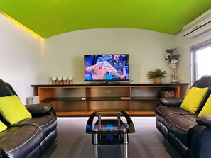 Een woonkamer met een televisie, twee leren banken met gele kussens, een glazen salontafel en een groen plafond.