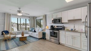 A modern kitchen with white cabinets, stainless steel appliances, and a dining area with a white sofa and wicker chair. - Sailfish Suites 1 - Waterfront & pet friendly! (Stuart)