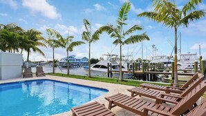 A swimming pool with lounge chairs, surrounded by palm trees and boats in the background. - Sailfish Suites 1 - Waterfront & pet friendly! (Stuart)