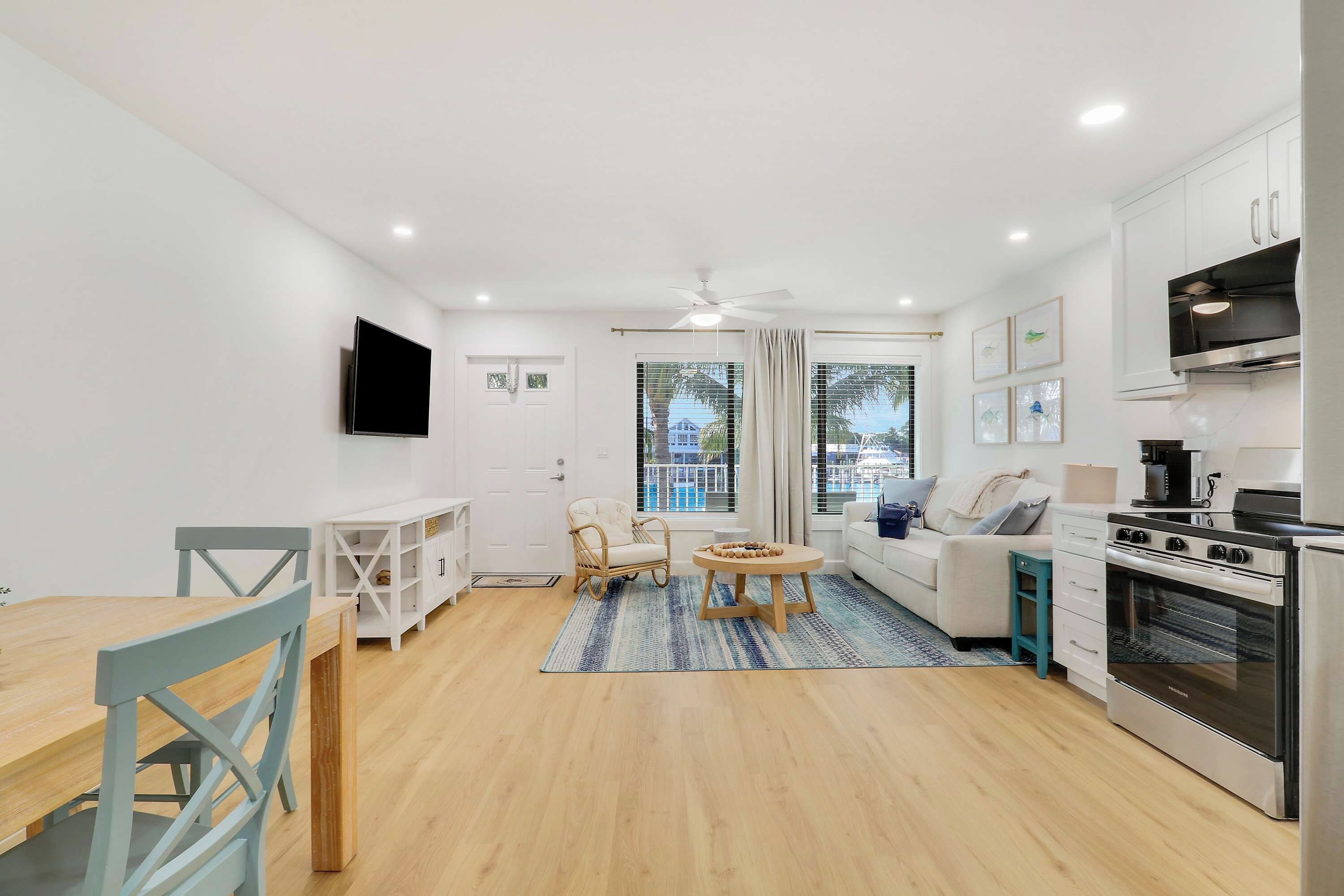 A modern living room with a dining area, a kitchen with a stove and oven, and a large window with a view of a pool.