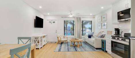 A modern living room with a dining area, a kitchen with a stove and oven, and a large window with a view of a pool.
