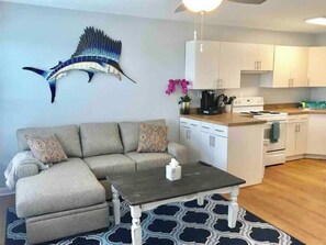 A living room with a sofa, a coffee table, a wall-mounted fish sculpture, a kitchen with white cabinets, and a patterned rug.