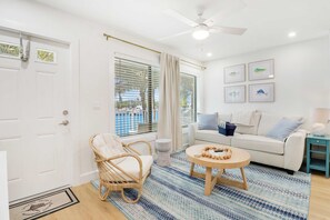 A room with a white door, a wicker chair, a white sofa, a round wooden table, and a ceiling fan.
