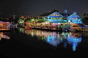 Une propriété située au bord de l’eau, avec une grande maison à plusieurs niveaux et dotée de balcons, un quai avec des bateaux, et une surface d’eau qui reflète l’environnement.