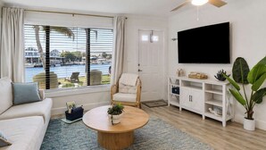Un soggiorno con un divano, un tavolino rotondo, una TV, una ventola a soffitto e vista su una distesa d'acqua attraverso ampie finestre.