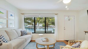 A room with a sofa, a round table, a wicker chair, a door, and a window with a view of a pool and palm trees. - Sailfish Suites 8 - Waterfront Lodging (Stuart)