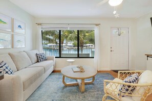 Une pièce comprenant un canapé, une table ronde, un fauteuil en osier, une porte et une fenêtre donnant sur une piscine et des palmiers.
