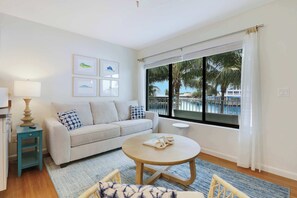 A modern living room with a sofa, a round coffee table, a small side table, and a large window with a view of palm trees and a pool.