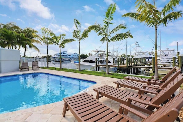 A swimming pool with lounge chairs, palm trees, and boats in the background.