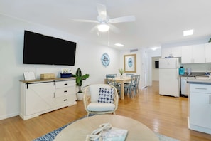 A modern living room with a flat-screen TV, a dining area with a table and chairs, a kitchen with white cabinets and appliances, and a ceiling fan.