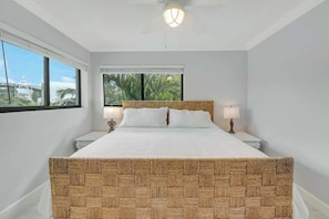 A bedroom with a large bed, two bedside tables, a ceiling fan, and a view of palm trees through the window. - Sailfish Suites - 12 - waterfront lodging (Stuart)