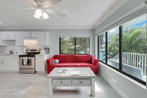 A modern kitchen with white cabinets, a red sofa, a white coffee table, and a large window with a view of palm trees. - Sailfish Suites - 12 - waterfront lodging (Stuart)