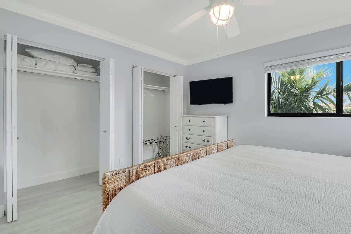 Quarto com cama, televisão na parede, armário e janela com vista para palmeiras.