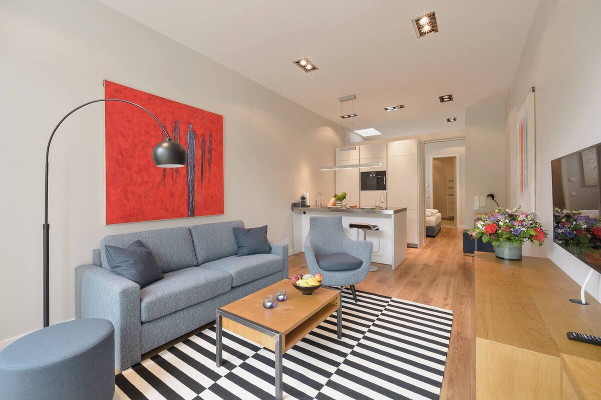 Ein modernes Wohnzimmer mit einer grauen Couch, einem Couchtisch und einem schwarz-weiß gestreiften Teppich. An der Wand hängt ein rotes abstraktes Gemälde, im Hintergrund ist eine Küchenzeile zu sehen.