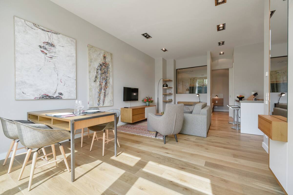 Ein modernes Wohnzimmer mit Holzboden, einem Essbereich mit Tisch und Stühlen, einem grauen Sofa und zwei abstrakten Bildern an der Wand.