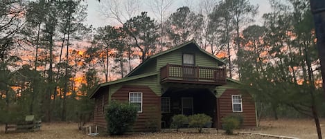 A rustic cabin with a front porch and a covered entryway, surrounded by trees and a dry ground.