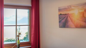 A room with a large window, red curtains, a vase with flowers, and a sunset painting on the wall.