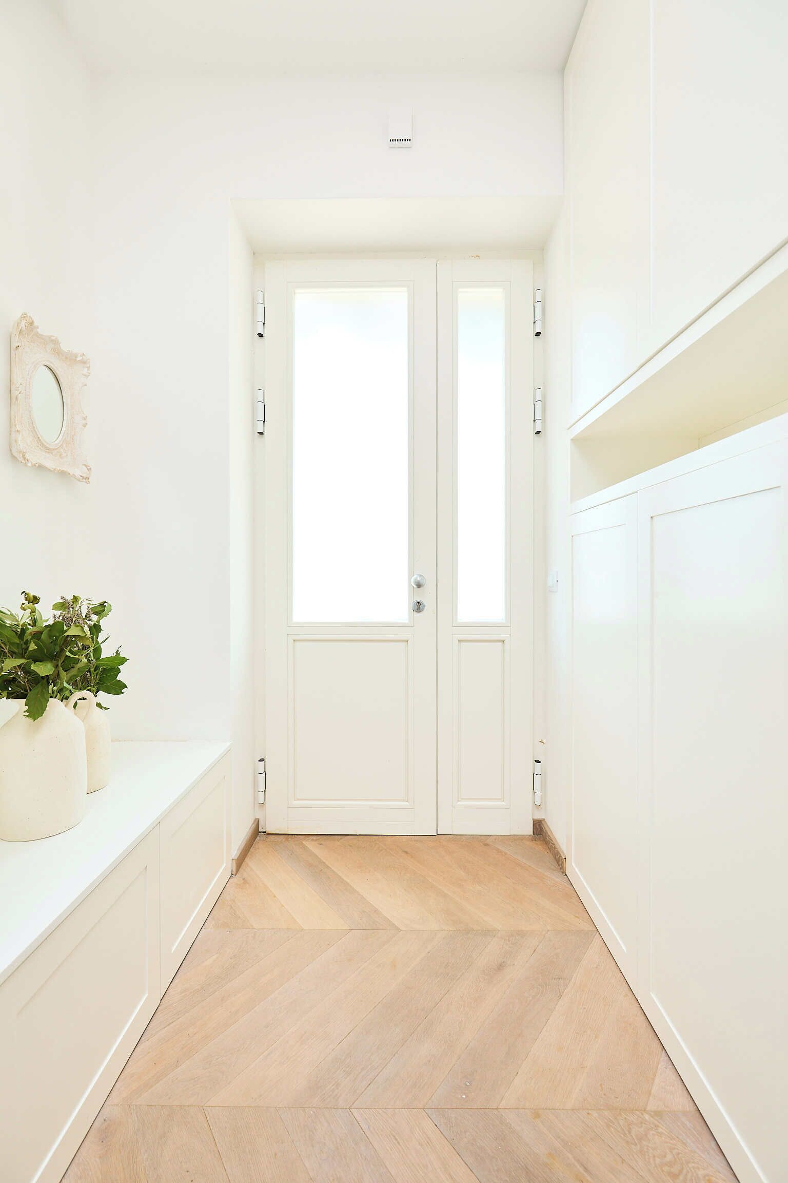 A hallway with a white door, a small plant on a white shelf, and a herringbone-patterned wooden floor.