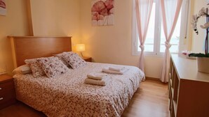 Una cama bien tendida con ropa de cama floral, una lámpara de noche y una ventana con cortinas translúcidas.
