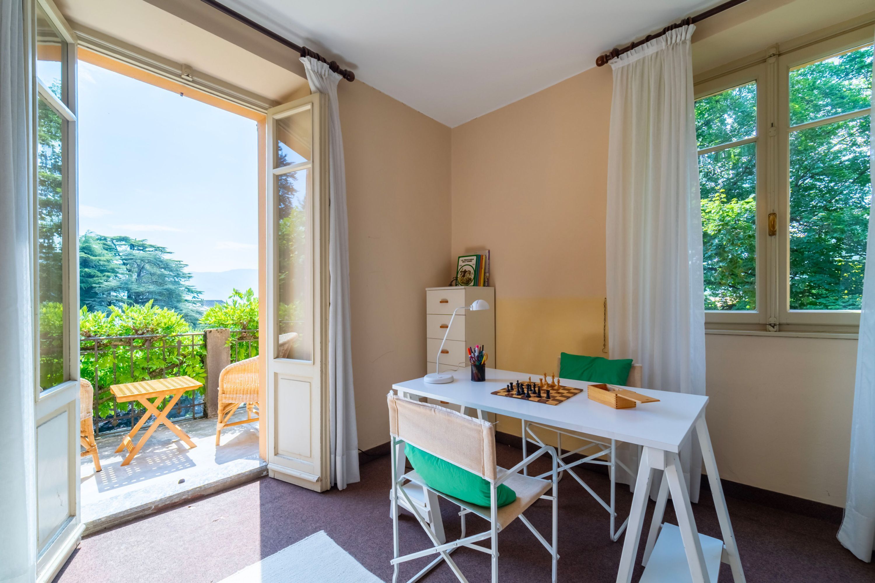 Une pièce avec un balcon, un bureau blanc, une chaise, une lampe et un jeu d’échecs.