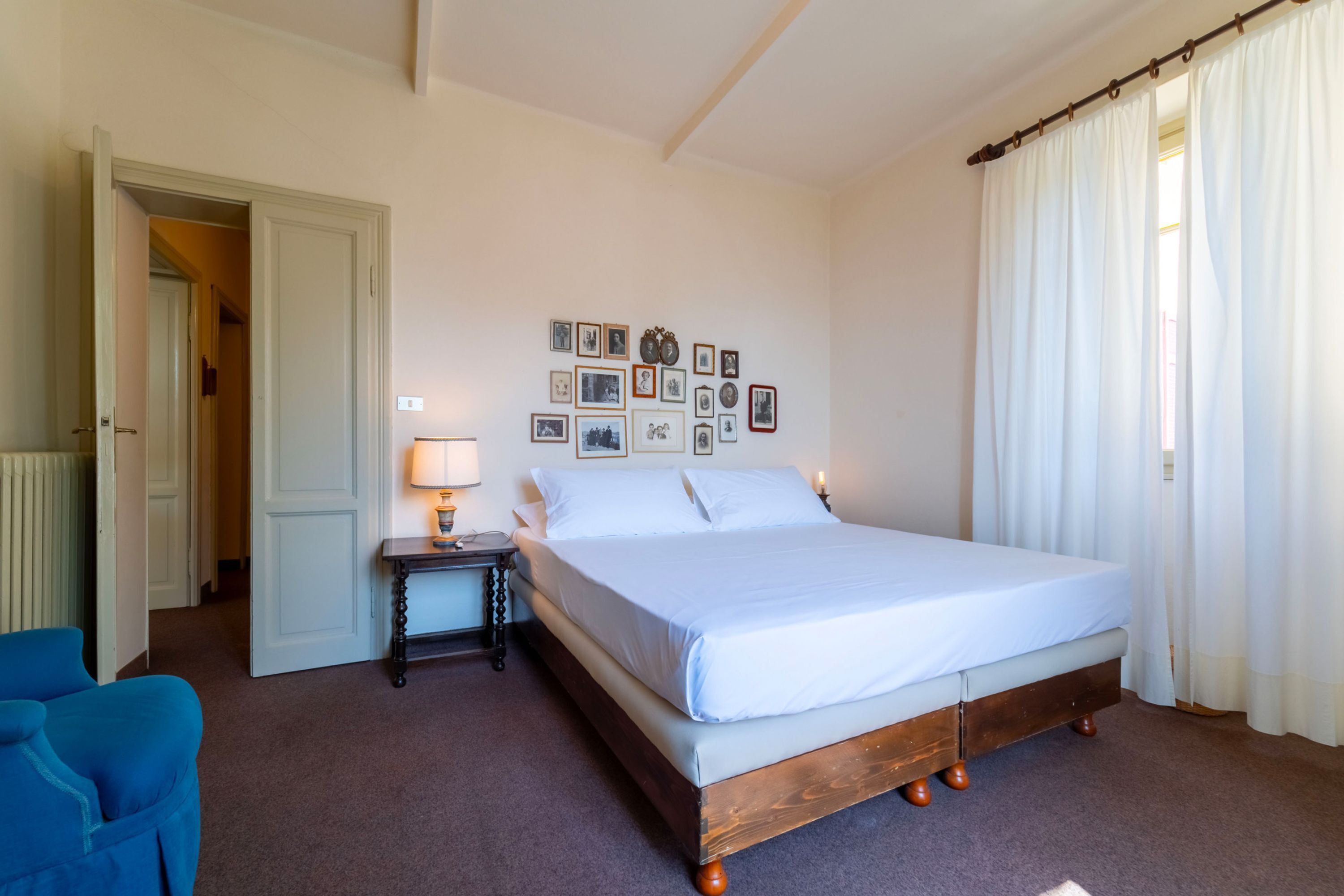 A bedroom with a bed, a nightstand, a lamp, a blue chair, and a wall with framed pictures.
