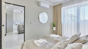 Une chambre moderne avec un grand lit, un miroir rond, une plante en pot et une vue sur l’extérieur à travers des rideaux légers.